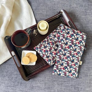 May include: A dark wooden serving tray holds a mug of coffee, a small plate with cookies, a lit candle, and a floral-patterned neck wrap. The neck wrap features a navy background with red, white, and blue flowers.