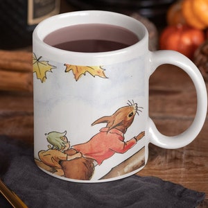 May include: White ceramic mug with a handle, featuring an illustration of a squirrel carrying a child on its back. The mug is filled with a dark liquid and is sitting on a wooden table with a grey cloth napkin and a gold spoon.
