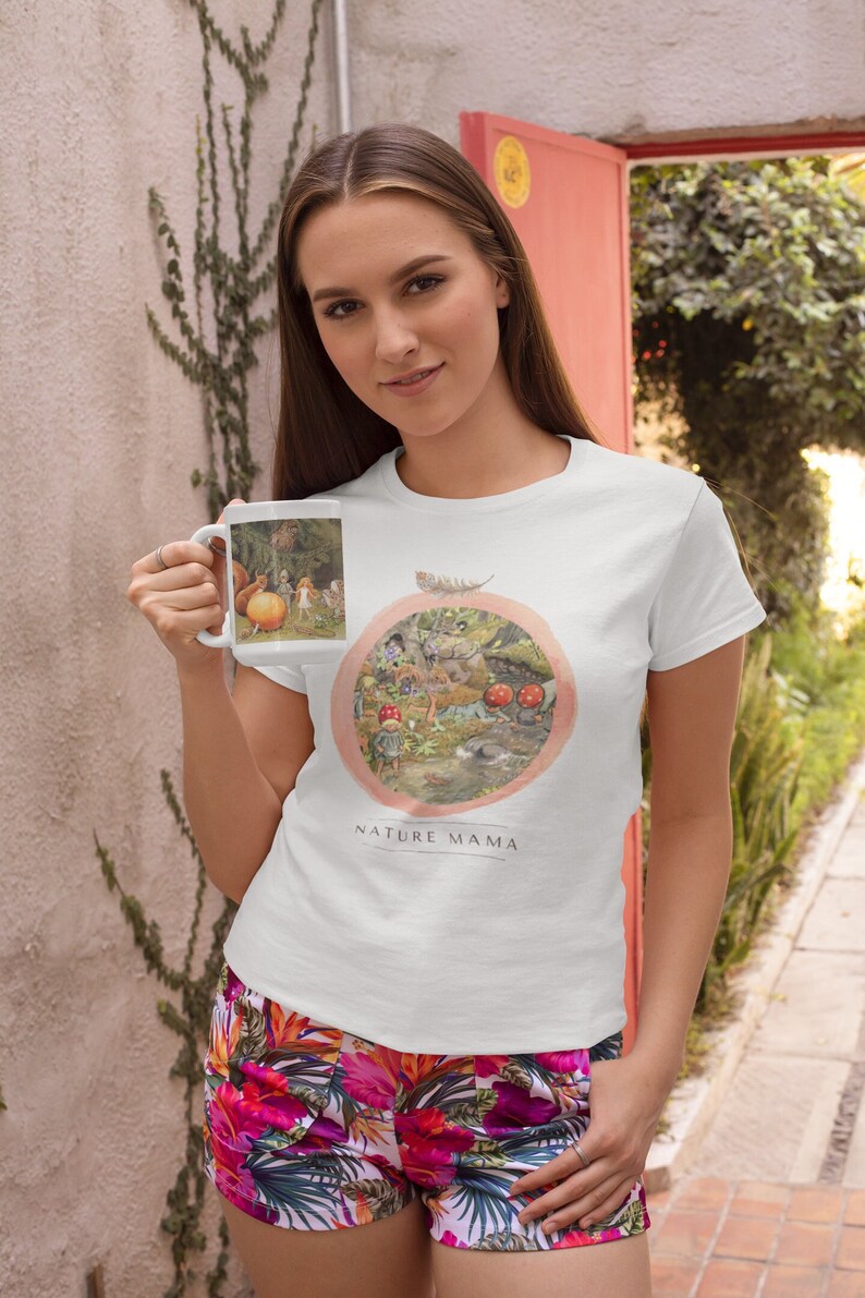 May include: White t-shirt with a nature-themed illustration of a forest scene with mushrooms, animals, and a feather. The text "NATURE MAMA" is printed below the illustration.