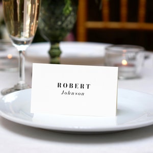 Pu&ograve; includere: Un segnaposto bianco con testo nero che recita "ROBERT Johnson" su un piatto bianco con uno sfondo di un bicchiere di champagne.