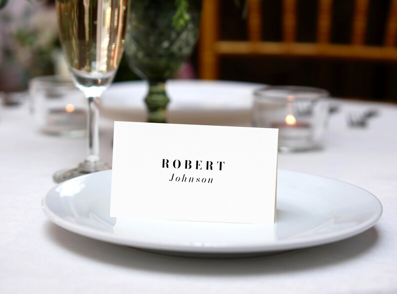 Pu&ograve; includere: Un segnaposto bianco con testo nero che recita "ROBERT Johnson" su un piatto bianco con uno sfondo di un bicchiere di champagne.