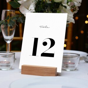 Pu&ograve; includere: Un biglietto bianco con testo nero che dice "Tavolo 12" su un supporto in legno. Il biglietto &egrave; su una tovaglia bianca con candele e bicchieri.