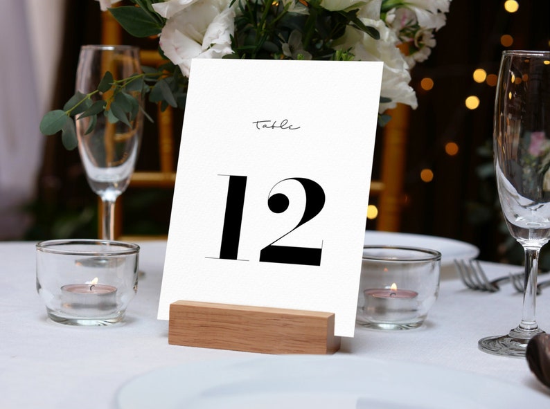 Pu&ograve; includere: Un biglietto bianco con testo nero che dice "Tavolo 12" su un supporto in legno. Il biglietto &egrave; su una tovaglia bianca con candele e bicchieri.