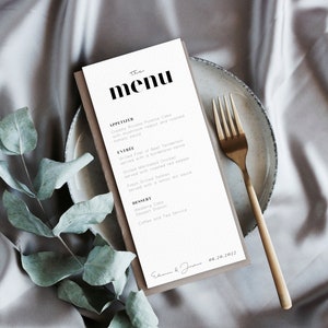 Pu&ograve; includere: Un menu bianco con testo nero che elenca gli antipasti, i piatti principali e i dessert per un ricevimento di nozze. Il menu &egrave; su un sottopiatto marrone con una forchetta dorata su una superficie beige. La data "08.20.2022" &egrave; scritta in fondo al menu.