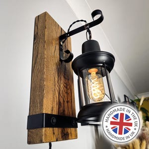 May include: A wall-mounted light fixture with a black metal lantern design and a visible Edison bulb. It's attached to a weathered wooden beam. The fixture has a black cord and is fixed to the wall. The light is handmade in the UK.