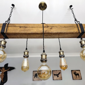 May include: A rustic wooden beam chandelier with five Edison bulb lights hanging from black cords. The beam is stained a dark brown and has black metal brackets on each end. The bulbs are clear glass with a gold finish.