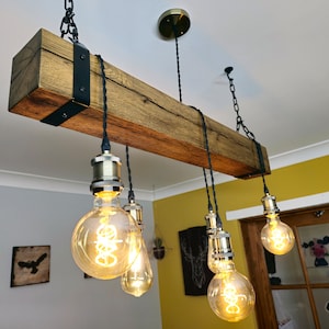 May include: A rustic wooden beam chandelier with six Edison bulb lights hanging from black cords and chains. The beam is stained a dark brown and has black metal brackets on each end. The bulbs are all lit and casting a warm glow.