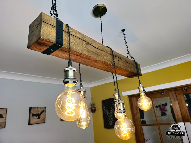 May include: A rustic wooden beam chandelier with six Edison bulb lights hanging from black cords and chains. The beam is stained a dark brown and has black metal brackets on each end. The bulbs are all lit and casting a warm glow.