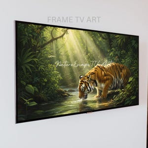 May include: A framed digital art piece depicting a tiger drinking from a stream in a lush jungle setting. The artwork features vibrant greens and browns, with sunlight streaming through the canopy. The text "FRAME TV ART" is at the top.