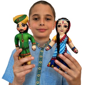 May include: A young person holding two handmade fabric dolls. The doll on the left is wearing a green tunic, yellow turban, and a brown belt. The doll on the right is wearing a blue and red sari with a yellow top and a black braid.