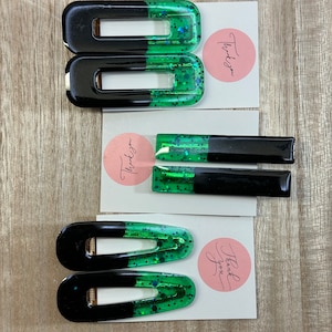 May include: Three pairs of black and green hair clips with glitter. The clips are in the shape of a rectangle with rounded edges. The top half of each clip is green with glitter and the bottom half is black.