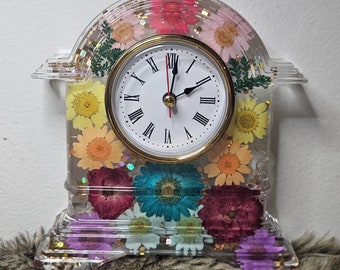 May include: A white clock with Roman numerals and a clear face is surrounded by pressed flowers in a clear resin clock base.