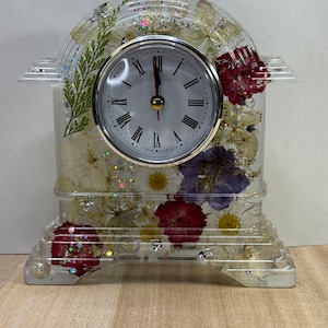 Może przedstawiać: Przezroczysty zegar z żywicy z białą tarczą i czarnymi rzymskimi cyframi godzinowymi. Zegar jest osadzony w przezroczystej żywicy z suszonymi kwiatami i brokatem.
