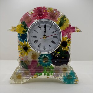May include: A decorative clock with a clear resin body, showcasing a variety of colorful dried flowers. The clock face is white with black Roman numerals and a silver bezel. The clock is approximately 6 inches tall.