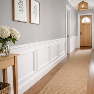 Puede incluir: Un pasillo con paredes gris claro, paneles blancos y una alfombra corredora tejida. Dos grabados botánicos enmarcados cuelgan de la pared. Una mesa de madera exhibe un jarrón con flores blancas. Una lámpara colgante tejida cuelga del techo.