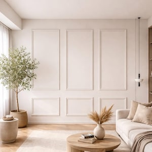 May include: A living room interior featuring a wall with white rectangular moulding panels, a potted olive tree in a woven basket, a round wooden coffee table with books, a beige sofa with cushions, and a modern pendant light fixture.