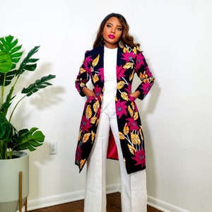 May include: A long black floral print coat with pink and yellow flowers. The coat has a collar and long sleeves. The model is wearing white trousers.