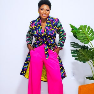 May include: A woman wearing a long, colorful patterned coat with a black belt and bright pink pants. The coat has a geometric design with yellow, green, blue, and pink accents.