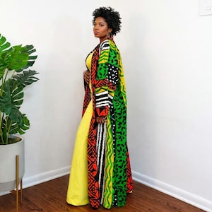 May include: A long, patterned duster coat with a vibrant mix of colors, including green, red, black, and yellow. The coat is worn over a yellow jumpsuit. The coat's design features geometric and floral patterns. The outfit is set against a white wall and a potted plant.