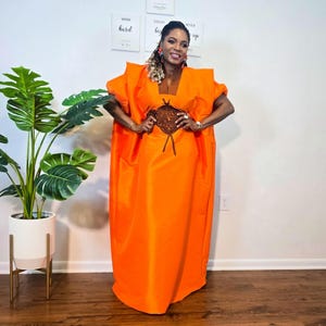 May include: Bright orange caftan dress with a brown embroidered design at the waist. The dress has short sleeves and a loose, flowing fit. The model is wearing the dress in a well-lit room.