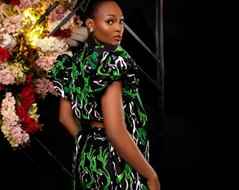 May include: A black two-piece outfit with a green and white leaf design. The top has short, puffed sleeves and a cropped style. The matching shorts complete the ensemble. The outfit is set against a backdrop of flowers.