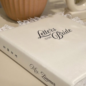 May include: A white book titled "Letters to the Bride" with black script, trimmed with lace. The spine displays the year "2025" and the name "Mrs. Zimmer". The book is on a light surface, with a brown vase and white decorative object in the background.