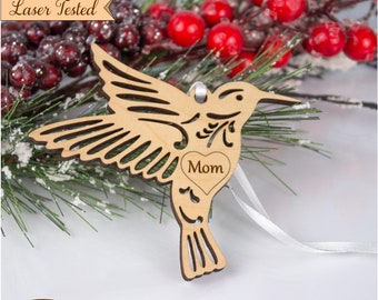 Può includere: Ciondolo a forma di colibrì in legno tagliato al laser con un ritaglio a forma di cuore che dice "Mamma". Il ciondolo è appeso a un nastro bianco ed è circondato da bacche rosse.