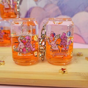 May include: Clear, can-shaped keychains filled with orange liquid. Each keychain features a cartoon design of two rabbits with the text "APPLE & HONEY" and "NATURAL". The keychains are connected by a silver chain.