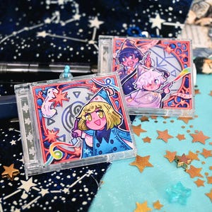 Może przedstawiać: Dwie kwadratowe, przezroczyste zawieszki akrylowe z kolorowymi ilustracjami w stylu anime przedstawiającymi postacie w czarodziejskich kapeluszach. Przednia zawieszka ukazuje blond postać w niebieskiej pelerynie, tylna dwie postacie z symbolami.
