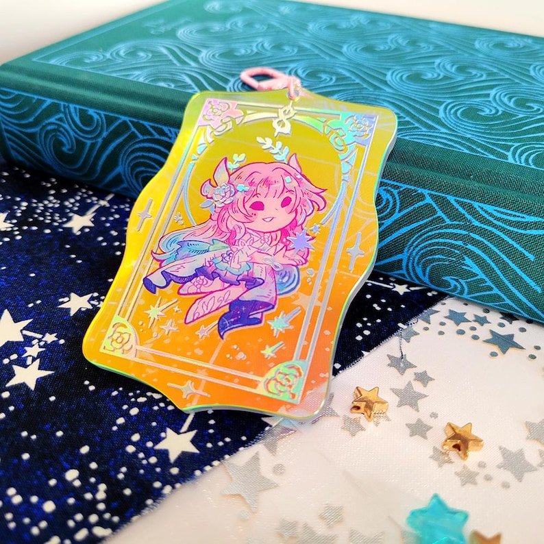 May include: A holographic charm featuring a cartoon character with pink hair and a floral headpiece. The charm has a yellow and orange gradient background with a decorative border. A pink string is attached to the top.