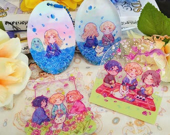 以下が含まれることがあります： 様々なアニメスタイルのアクリルチャームとキーホルダー。キーホルダーは楕円形で、様々な背景のキャラクターのイラストが描かれています。1つのチャームは、赤い毛布と白い猫がいるピクニックのシーンを描いています。