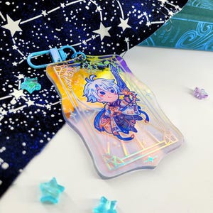 May include: A rectangular, iridescent acrylic keychain featuring a cartoon character with a sword. The character has blue hair and is wearing a blue and white outfit. The keychain has a blue clasp and is surrounded by star-shaped decorations.