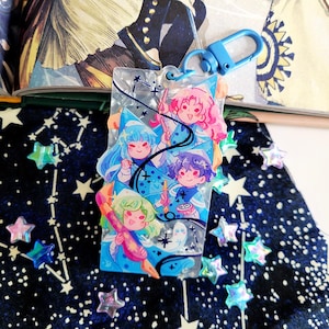 May include: A clear acrylic keychain featuring a colorful illustration of four anime-style characters with blue, pink, and green hair. The keychain has a blue clasp and is decorated with stars and other small details.