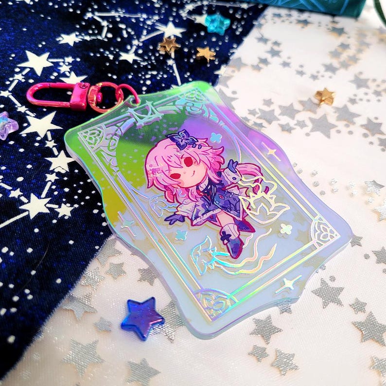 May include: A holographic acrylic keychain featuring a cartoon character with pink hair and a blue outfit. The keychain has a pink clasp and a decorative border with a green and purple gradient. The background includes star-shaped confetti.