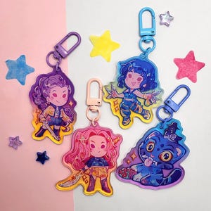 May include: Collection of colorful acrylic keychains featuring cartoon characters and a blue cat. Each keychain has a different character design with vibrant colors and attached to a metal clasp. Decorative star-shaped embellishments are scattered around the keychains.