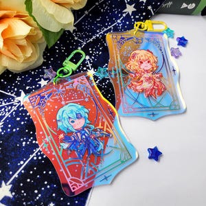 May include: Two acrylic keychains featuring anime-style illustrations of characters. One keychain has a red and orange background, the other has a blue background. Both have a gold-colored clasp and star-shaped charms. The keychains are on a starry background.