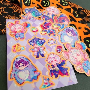 May include: A sheet of colorful stickers featuring cartoon characters in various costumes, including a shark, bunny, and vampire. The stickers are on a purple background with a diamond pattern. Halloween-themed fabric is in the background.