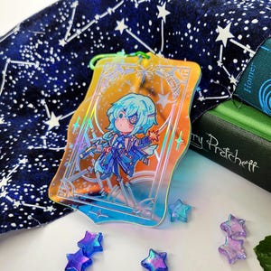 May include: A colorful acrylic charm featuring a cartoon character with blue hair and clothing, holding a star. The charm has a holographic effect and is surrounded by a decorative frame. Small star-shaped charms are scattered around the main charm.