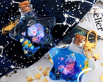 May include: Two star-shaped glass bottles with cork stoppers, each containing blue liquid, glitter, and a small illustrated character. Each bottle features a gold-toned metal keychain attachment. The bottles are filled with tiny star-shaped confetti.