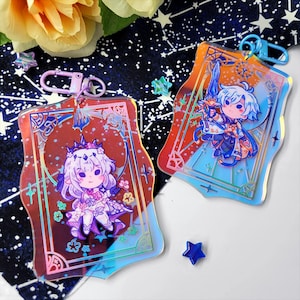 May include: Two holographic acrylic keychains featuring anime-style characters. One keychain has a character with a crown and floral accents, the other has a character holding a sword. Both have a decorative border and a metal clasp.