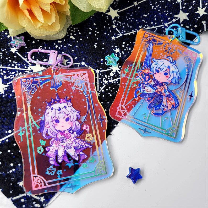 May include: Two holographic acrylic keychains featuring anime-style characters. One keychain has a character with a crown and floral accents, the other has a character holding a sword. Both have a decorative border and a metal clasp.