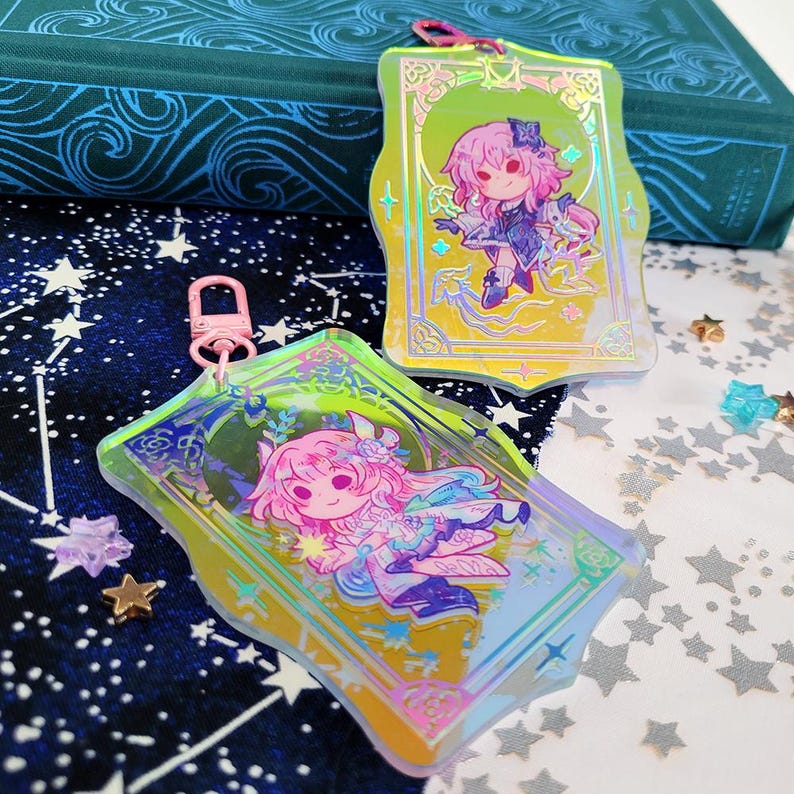 May include: Two acrylic keychains with cartoon illustrations of anime characters. The keychains have a holographic effect and are rectangular with rounded edges. One keychain has a pink clasp. The background is a starry pattern.