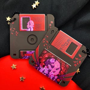 May include: Two dark brown floppy disk pendants with red and purple artwork. One pendant features a portrait, the other has a design with eyes and text. Both have red floral accents and are on black beaded chains.