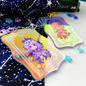 May include: Two iridescent acrylic keychains featuring anime-style illustrations. One keychain has a yellow background with a character in a floral dress. The other keychain has a similar design with a different character. Both have gold-colored frames and star-shaped accents.