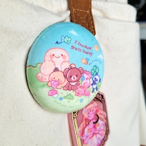 May include: A round, light blue button with the text "I touched grass today" and a cartoon illustration of various cute creatures in a grassy field. The button is attached to a beige bag with a brown strap.