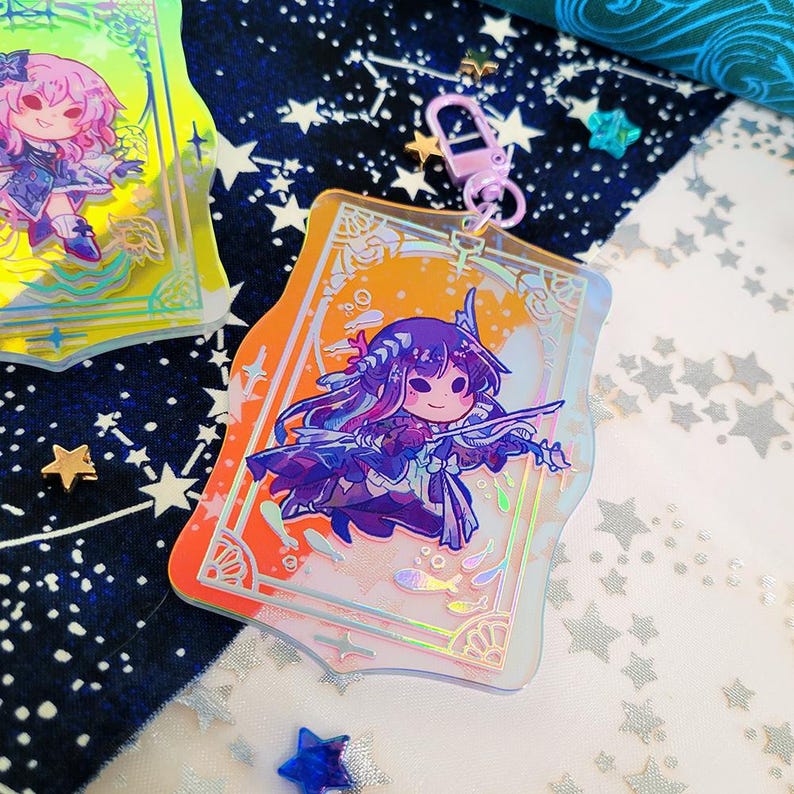 May include: A clear acrylic charm featuring a cartoon character with purple hair and a violin. The charm has an orange and yellow background with a decorative border and a pink clasp. The design includes fish and star details.