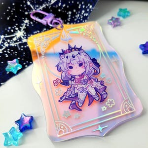May include: A clear acrylic charm featuring a cartoon character with purple hair and a crown, set against a pastel pink and blue background. The charm has a decorative border and a purple clasp.