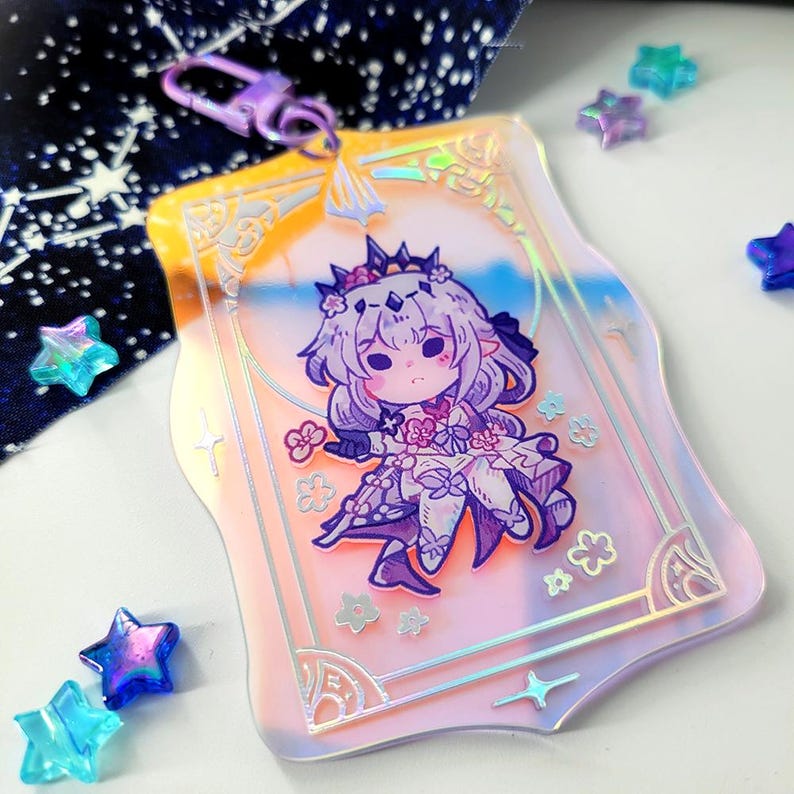 May include: A clear acrylic charm featuring a cartoon character with purple hair and a crown, set against a pastel pink and blue background. The charm has a decorative border and a purple clasp.