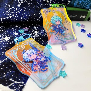 May include: Two acrylic keychains with cartoon character illustrations. The keychains have a rectangular shape with rounded edges and a gold and blue color scheme. One keychain has a blue clasp, and the other has a green clasp. Scattered around are star-shaped decorations.