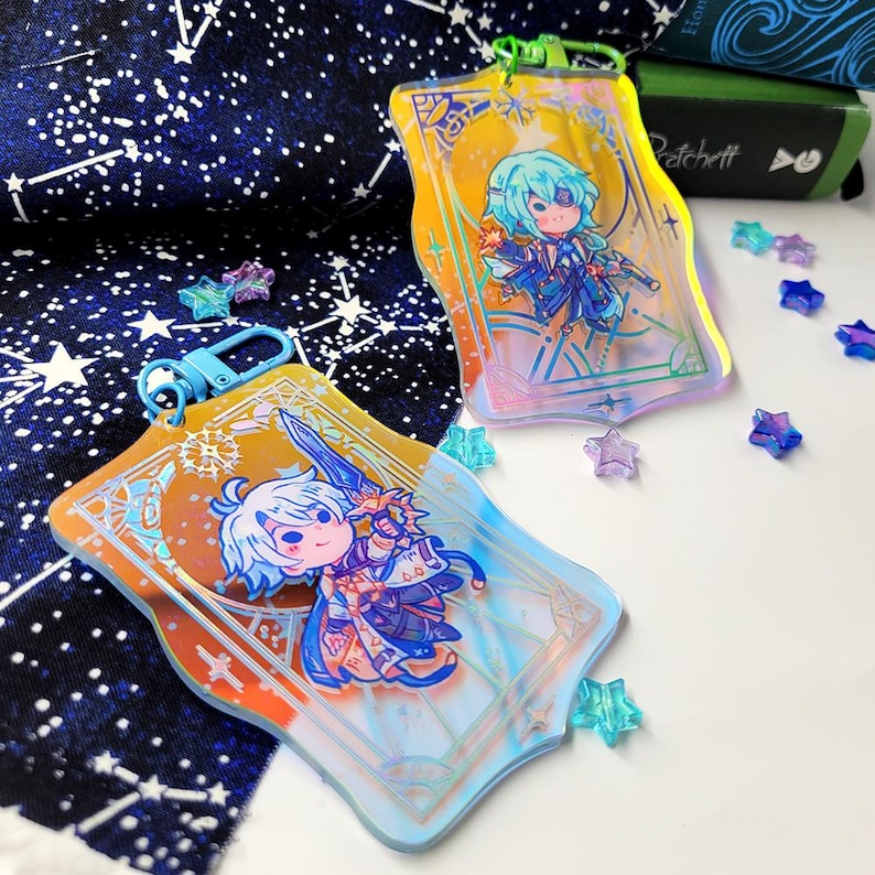May include: Two acrylic keychains with cartoon character illustrations. The keychains have a rectangular shape with rounded edges and a gold and blue color scheme. One keychain has a blue clasp, and the other has a green clasp. Scattered around are star-shaped decorations.